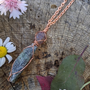 Peut inclure: Un collier pendentif artisanal en fil de cuivre avec une pierre verte et marron. La pierre est enveloppée de fil de cuivre et suspendue à une chaîne en cuivre. Le collier est présenté sur une surface en bois avec des fleurs et des feuilles.
