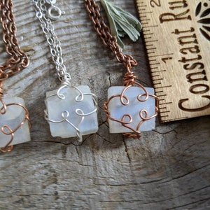 Puede incluir: Tres colgantes de alambre envueltos en plata y cobre con cristales de piedra lunar blanca. Los colgantes cuelgan de cadenas sobre una superficie de madera.