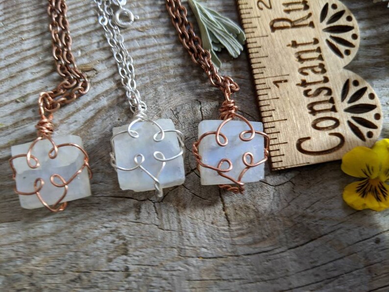 Puede incluir: Tres colgantes de alambre envueltos en plata y cobre con cristales de piedra lunar blanca. Los colgantes cuelgan de cadenas sobre una superficie de madera.