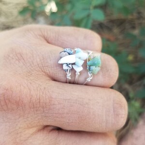 May include: Three silver rings with different colored stones. The rings are stacked on a finger. The stones are green, white, and black.