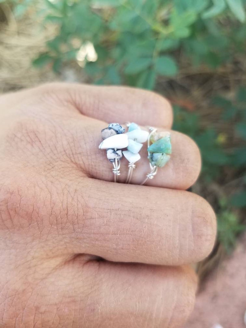 May include: Three silver rings with different colored stones. The rings are stacked on a finger. The stones are green, white, and black.