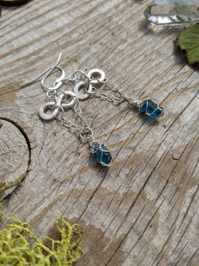 May include: Silver infinity symbol earrings with a dangling blue gemstone wrapped in silver wire.