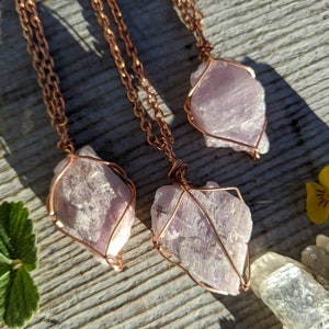 May include: Three copper wire wrapped pendants featuring raw amethyst crystals. The amethyst crystals are a light purple color and have a natural, rough texture. The copper wire is a warm, reddish-brown color.