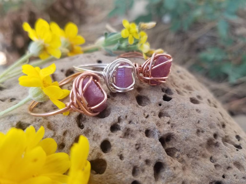 May include: Three wire-wrapped rings with pink and purple gemstones. The rings are made with copper and silver wire.