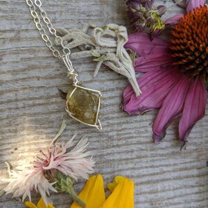 May include: A silver chain necklace with a wire-wrapped, yellow-green gemstone pendant. The pendant is a rough, natural stone with a unique shape. The necklace is displayed on a wooden surface with dried flowers.