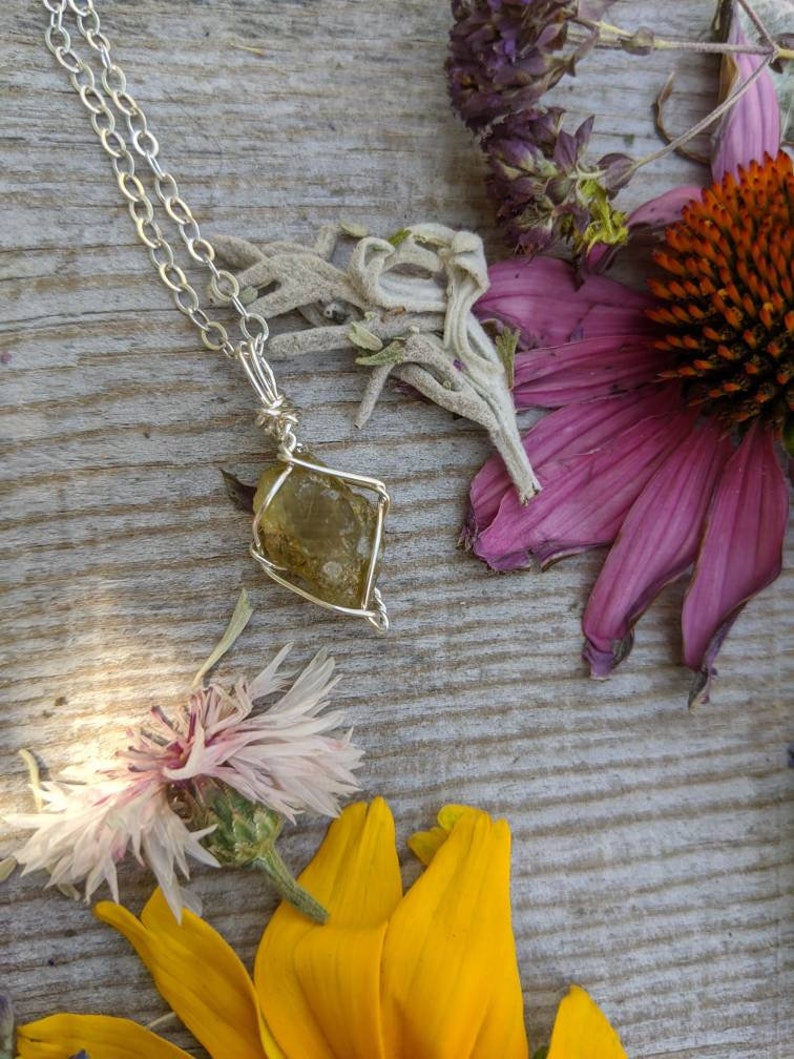 May include: A silver chain necklace with a wire-wrapped, yellow-green gemstone pendant. The pendant is a rough, natural stone with a unique shape. The necklace is displayed on a wooden surface with dried flowers.