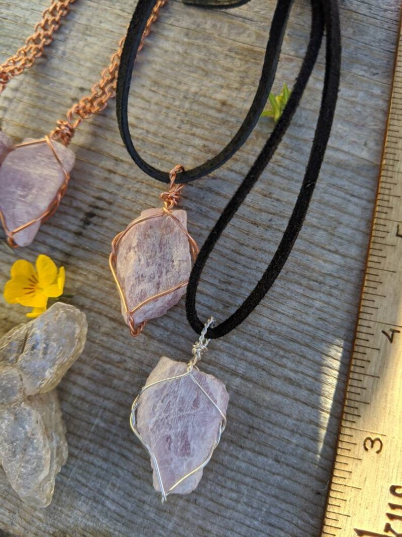 May include: Two necklaces with pink crystal pendants wrapped in wire. The necklaces are on a wooden surface with a ruler in the background. One necklace has a copper wire wrap and the other has a silver wire wrap.