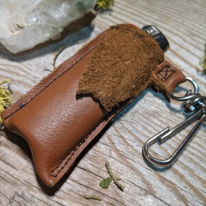 May include: A brown leather lighter case with a suede flap and a silver metal keychain. The case is stitched with black thread and has a small metal loop for attaching to a keychain.