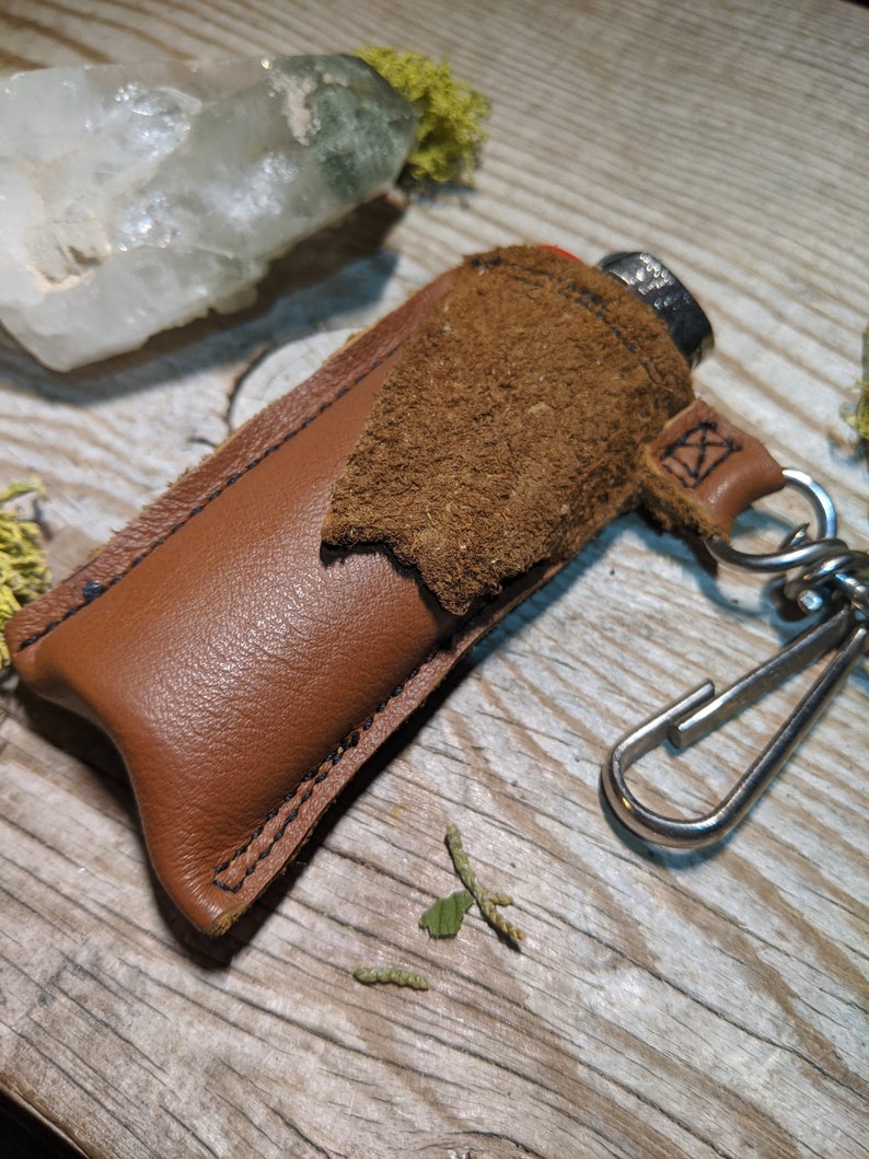 May include: A brown leather lighter case with a suede flap and a silver metal keychain. The case is stitched with black thread and has a small metal loop for attaching to a keychain.