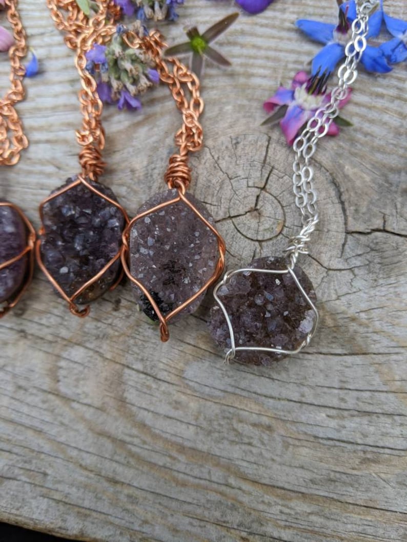May include: Three amethyst crystal necklaces with copper and silver wire wrapping. The crystals are a deep purple color and have a rough, natural texture. The necklaces are displayed on a wooden surface.