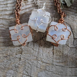 Puede incluir: Tres collares con colgantes de piedra lunar envueltos en alambre. Los colgantes son de forma cuadrada y tienen un acabado blanco iridiscente. Los collares est&aacute;n sobre una superficie de madera.
