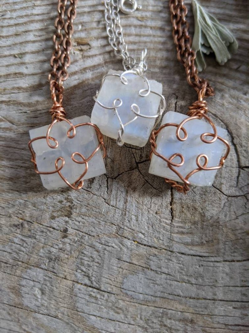 Puede incluir: Tres collares con colgantes de piedra lunar envueltos en alambre. Los colgantes son de forma cuadrada y tienen un acabado blanco iridiscente. Los collares est&aacute;n sobre una superficie de madera.