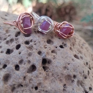 May include: Three rings with pink gemstones wrapped in copper wire. The rings are on a textured brown surface.