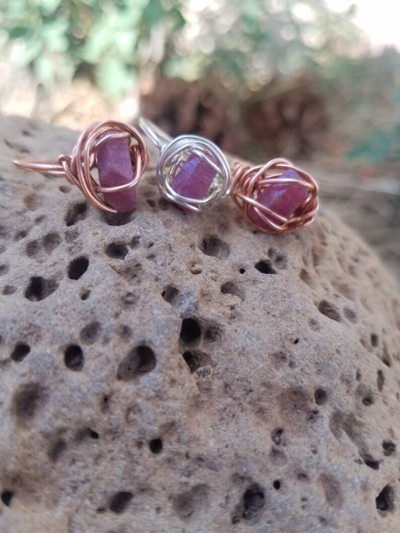 May include: Three rings with pink gemstones wrapped in copper wire. The rings are on a textured brown surface.