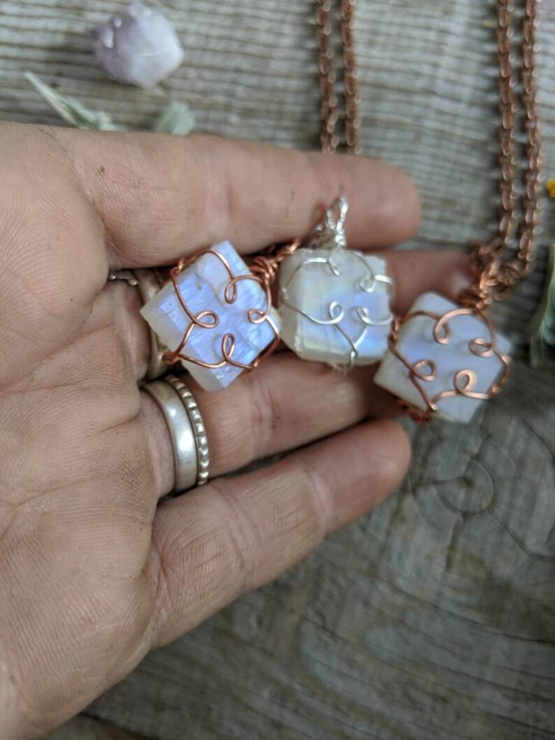 Puede incluir: Tres colgantes de piedra lunar iridiscente envueltos en alambre de cobre, cada uno con un dise&ntilde;o &uacute;nico. Los colgantes cuelgan de una cadena de cobre.