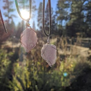 May include: Two pink crystal pendants on black leather cords. The crystals are wrapped in silver wire and hang from a natural setting.