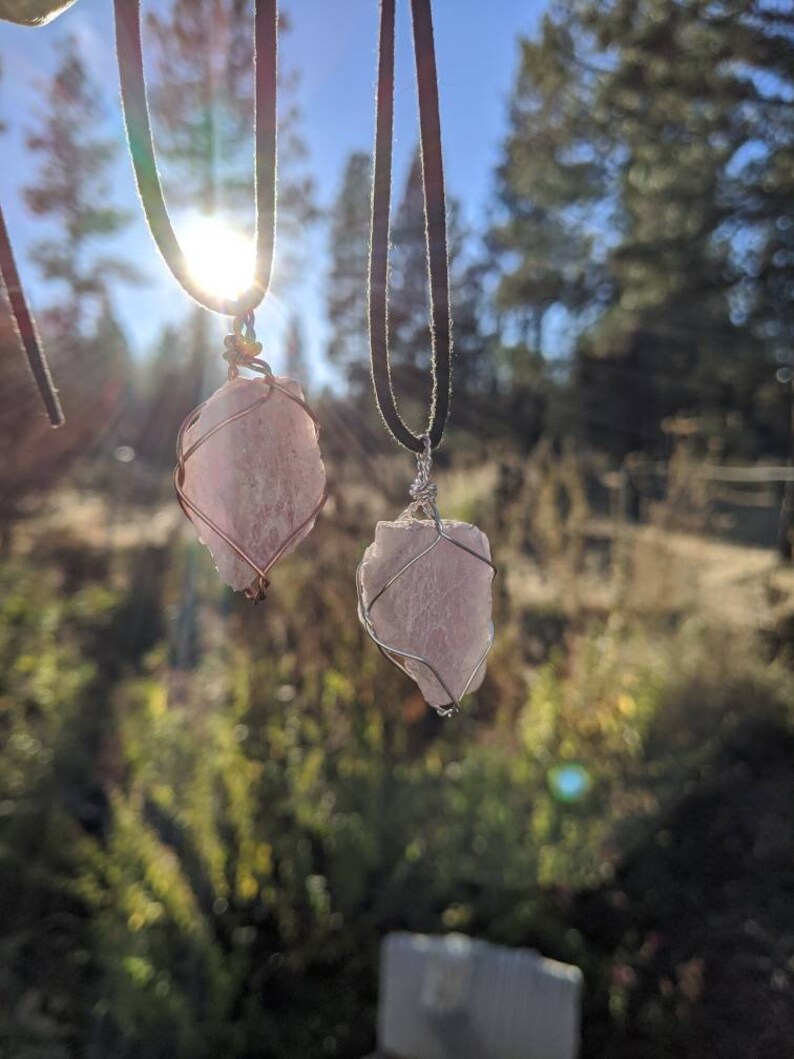 May include: Two pink crystal pendants on black leather cords. The crystals are wrapped in silver wire and hang from a natural setting.