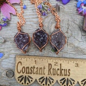 May include: Three amethyst crystal pendants wrapped in copper wire, hanging from copper chains. The pendants are displayed on a wooden surface with a ruler that reads "Constant Ruckus".
