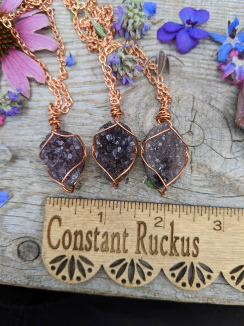 May include: Three amethyst crystal pendants wrapped in copper wire, hanging from copper chains. The pendants are displayed on a wooden surface with a ruler that reads "Constant Ruckus".