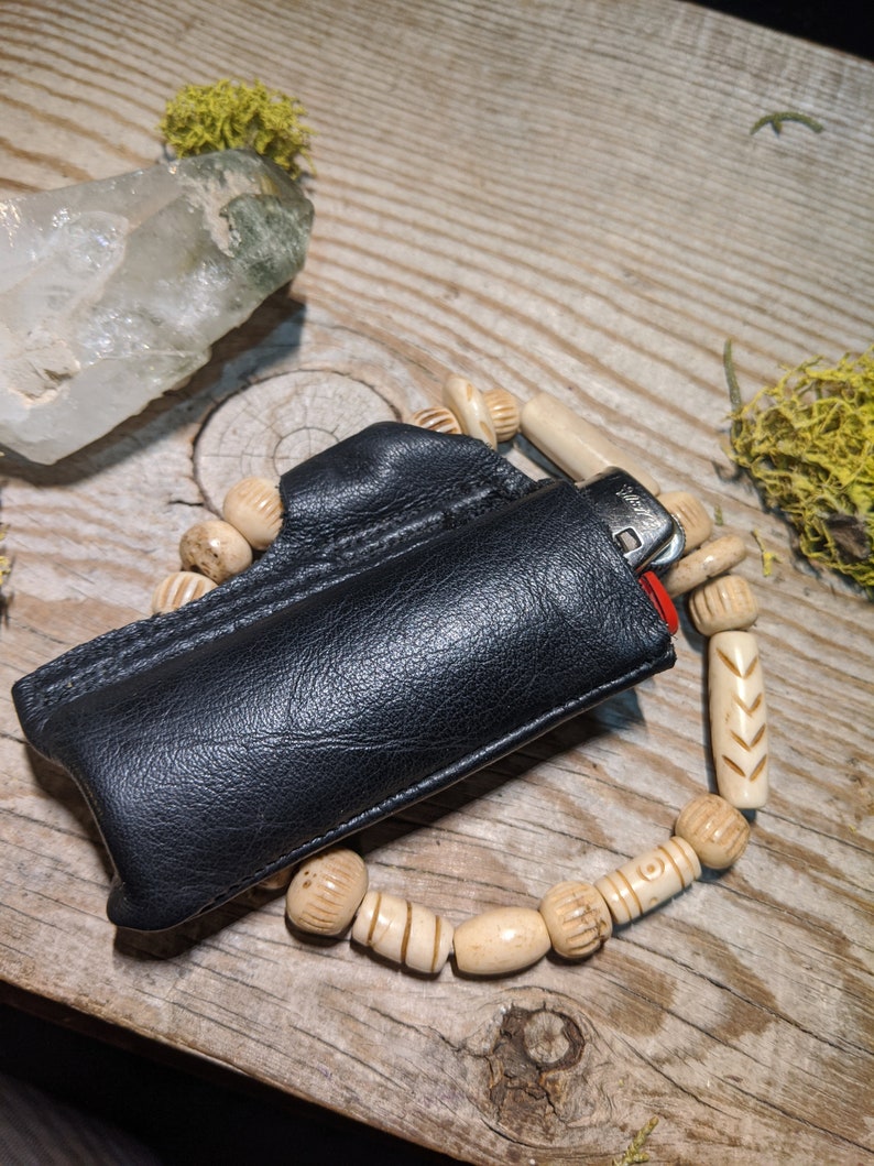 Puede incluir: Una funda de cuero negro para encendedor con un encendedor plateado en su interior. La funda est&aacute; decorada con una cadena de cuentas de madera blancas.