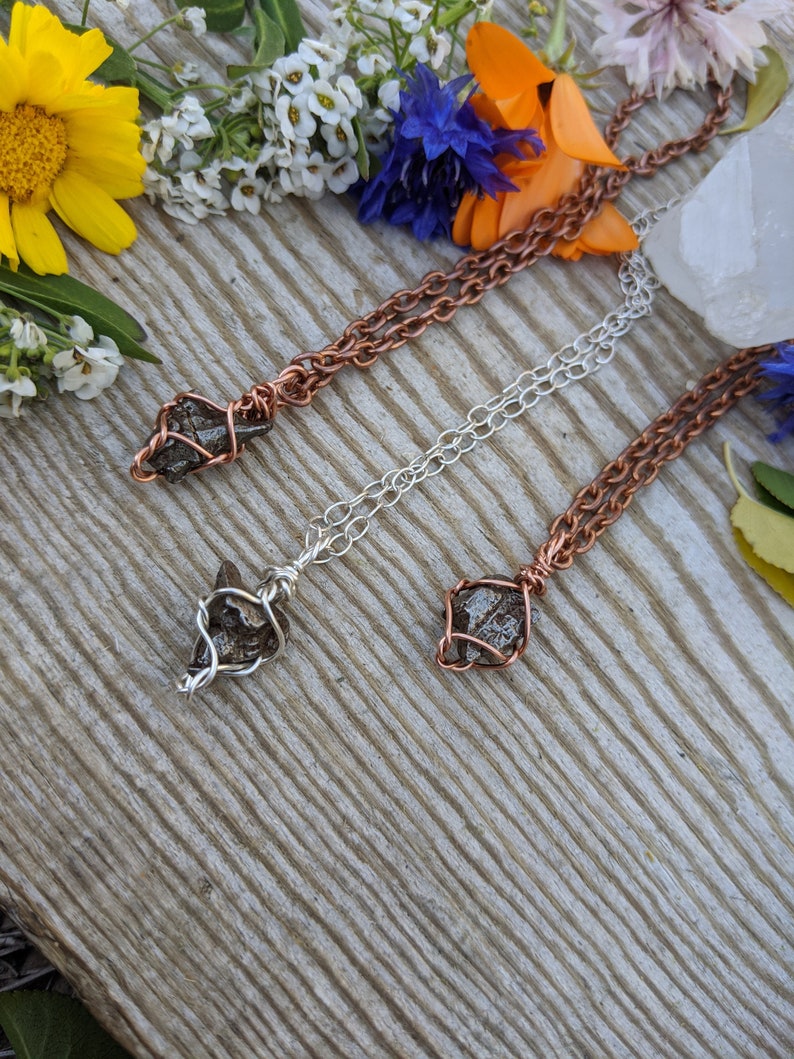 Puede incluir: Tres collares con cadenas de diferentes colores y colgantes de cristal envueltos en alambre. Las cadenas son de cobre, plata y cobre. Los cristales son de color gris oscuro y tienen una textura rugosa y natural.
