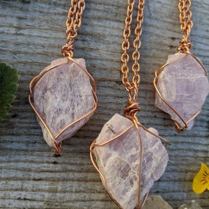 May include: Three pink crystal pendants wrapped in copper wire, hanging from copper chains. The crystals are rough and natural, with a light pink color. The pendants are displayed on a wooden surface.