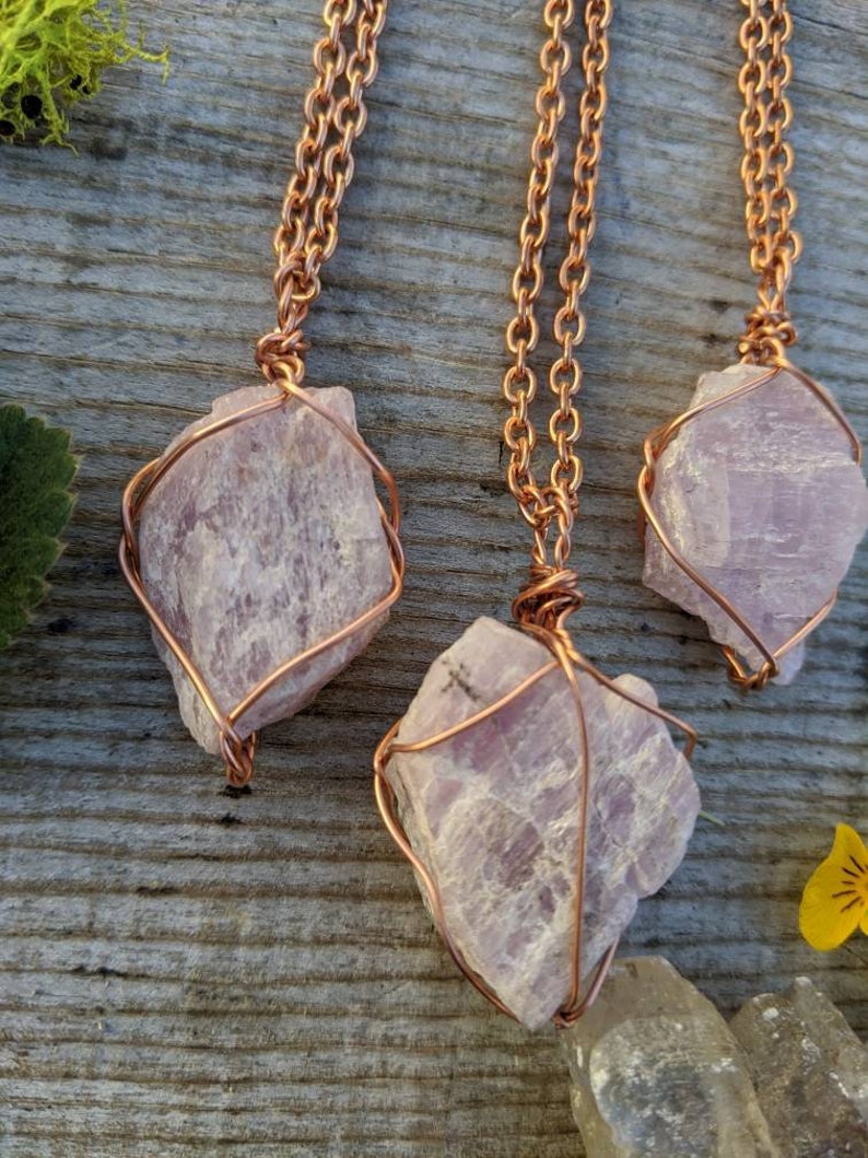 May include: Three pink crystal pendants wrapped in copper wire, hanging from copper chains. The crystals are rough and natural, with a light pink color. The pendants are displayed on a wooden surface.