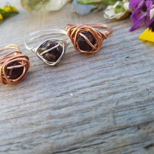May include: Three wire-wrapped rings with dark red gemstone beads. The rings are made with copper, silver, and gold wire.