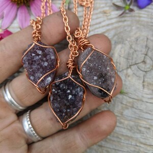 May include: Three amethyst crystal pendants wrapped in copper wire, hanging from copper chains. The pendants are different sizes and shapes, and the amethyst crystals are a deep purple color.