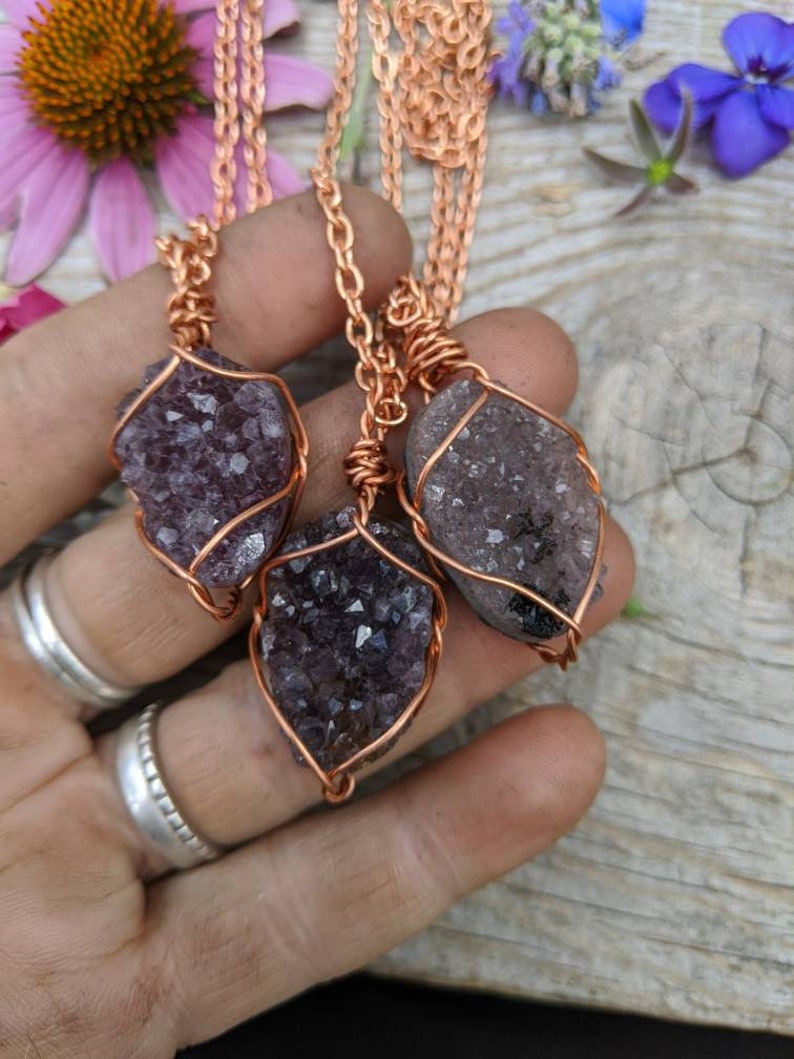 May include: Three amethyst crystal pendants wrapped in copper wire, hanging from copper chains. The pendants are different sizes and shapes, and the amethyst crystals are a deep purple color.