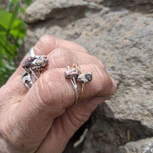 May include: Two rings with a wire design and a small, square, pink and black stone. The rings are on a person's finger.