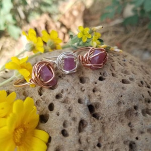 May include: Three wire-wrapped rings with pink and purple gemstones. The rings are on a brown, textured rock.