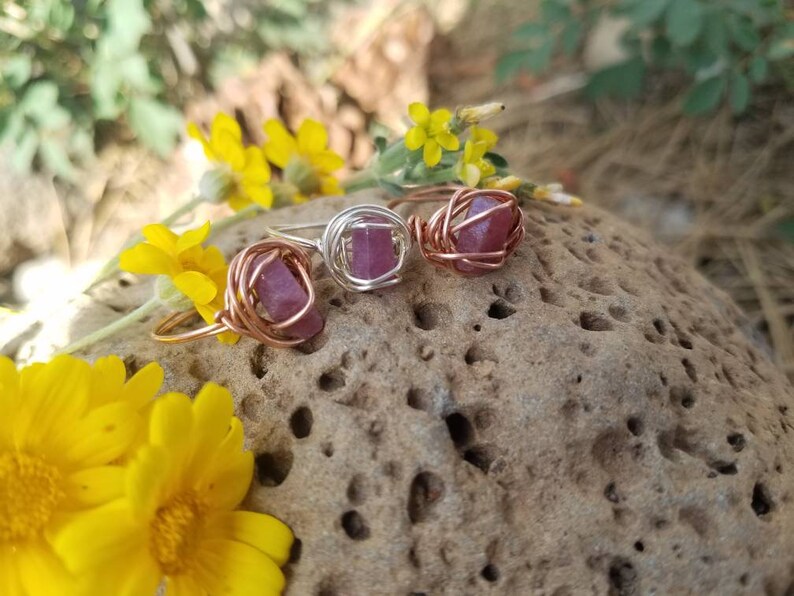 May include: Three wire-wrapped rings with pink and purple gemstones. The rings are on a brown, textured rock.