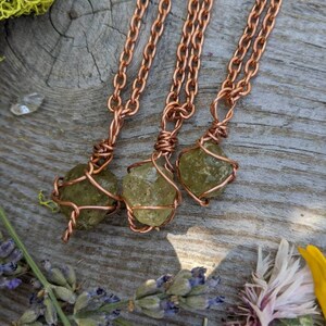 May include: Three copper wire wrapped green peridot stone pendants on copper chains. The pendants are hanging on a wooden surface with green moss and flowers.