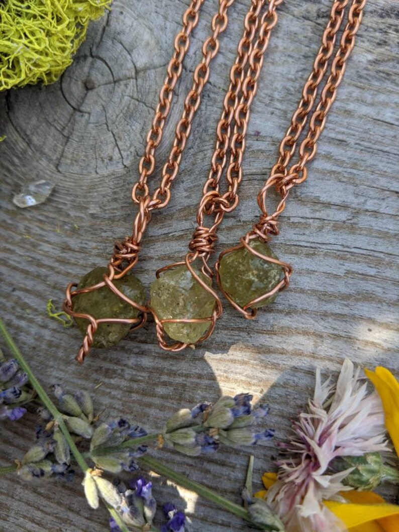 May include: Three copper wire wrapped green peridot stone pendants on copper chains. The pendants are hanging on a wooden surface with green moss and flowers.