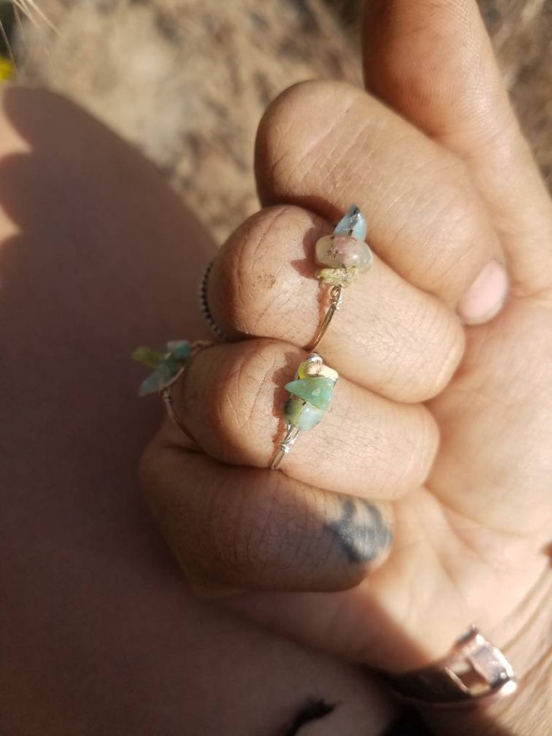 May include: Two rings with small green and blue stones wrapped in wire. The rings are on a person's finger.