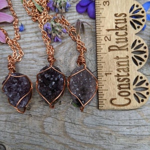 May include: Three amethyst crystal pendants wrapped in copper wire, hanging from copper chains. The pendants are displayed on a wooden surface with a ruler labeled "Constant Ruckus" in the background.