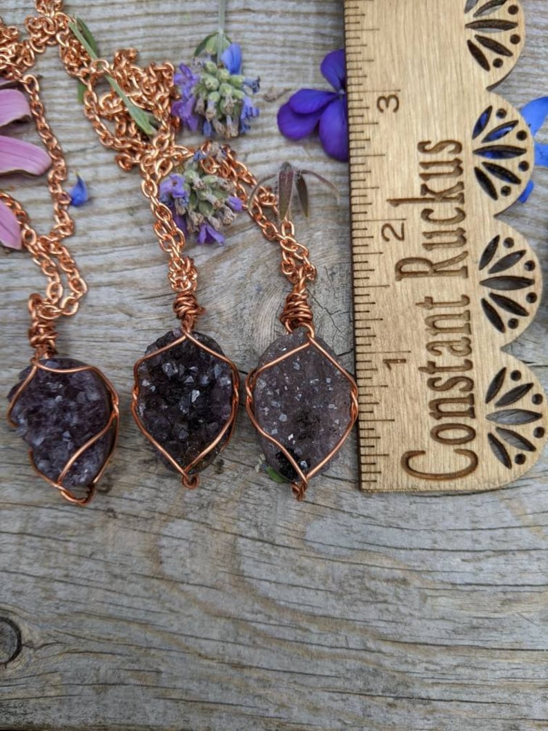 May include: Three amethyst crystal pendants wrapped in copper wire, hanging from copper chains. The pendants are displayed on a wooden surface with a ruler labeled "Constant Ruckus" in the background.