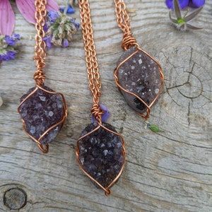 May include: Three amethyst crystal pendants wrapped in copper wire, hanging from copper chains. The pendants are on a wooden surface with purple flowers.