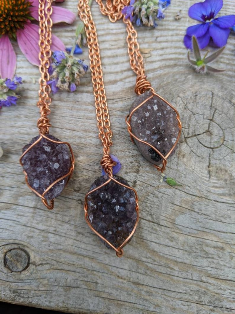 May include: Three amethyst crystal pendants wrapped in copper wire, hanging from copper chains. The pendants are on a wooden surface with purple flowers.