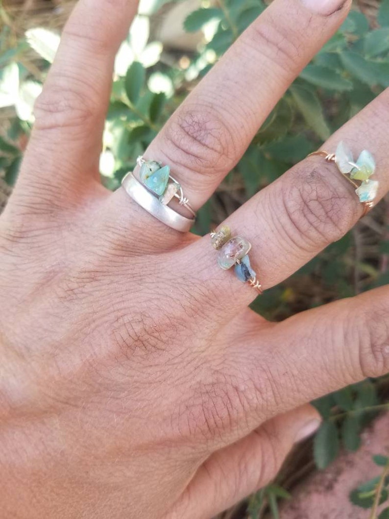 May include: Two rings with wire wrapped around them and small green and blue stones. The rings are on a person's hand.