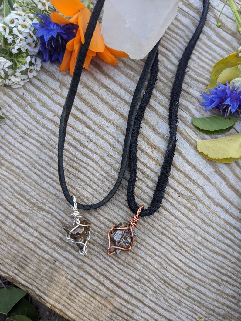 Puede incluir: Dos collares con piedras gris oscuro envueltas en alambre. Un collar tiene un envoltorio de alambre plateado y el otro tiene un envoltorio de alambre de cobre. Ambos collares est&aacute;n en cordones de gamuza negra.