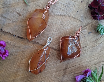 May include: Three gemstone pendants with wire wrapping, displayed on a wooden surface with flowers. The pendants feature orange-brown stones in various shapes: a large, irregular stone on a copper chain, a teardrop shape with silver wire, and a square stone with copper wire.