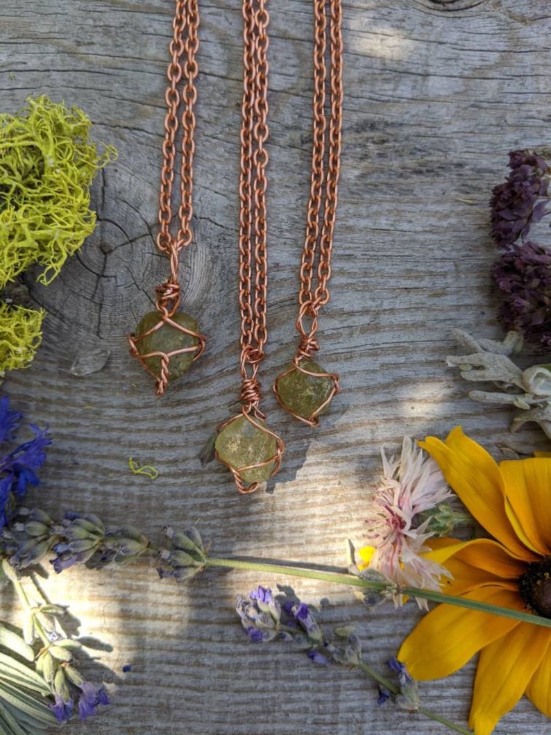 May include: Three copper chain necklaces with green gemstone pendants wrapped in copper wire. The necklaces are displayed on a wooden surface with flowers.