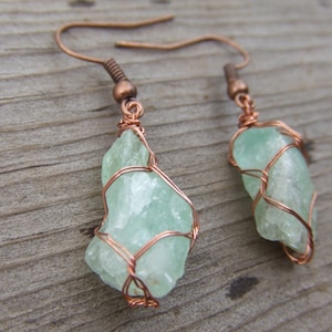 May include: A pair of copper wire wrapped earrings featuring light green, translucent, raw crystal stones.