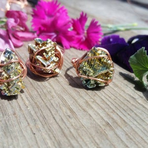 Może przedstawiać: Trzy pierścionki z miedzianym drutem owiniętym wokół tęczowych kryształów pirytu. Pierścionki leżą na drewnianej powierzchni z różowymi i fioletowymi kwiatami.