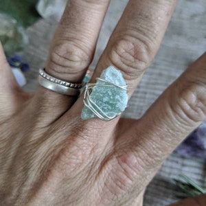 May include: A silver wire wrapped ring featuring a light green, rough-cut gemstone. The ring is on a person's finger.