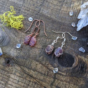 May include: A pair of earrings featuring small, irregular shaped, purple and pink stones wrapped in copper and silver wire. The earrings are on a wooden surface with small clear crystals scattered around them.