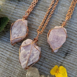 May include: Three delicate necklaces with pink crystal pendants wrapped in copper wire. The necklaces are displayed on a wooden surface with a yellow flower and a brown leaf.