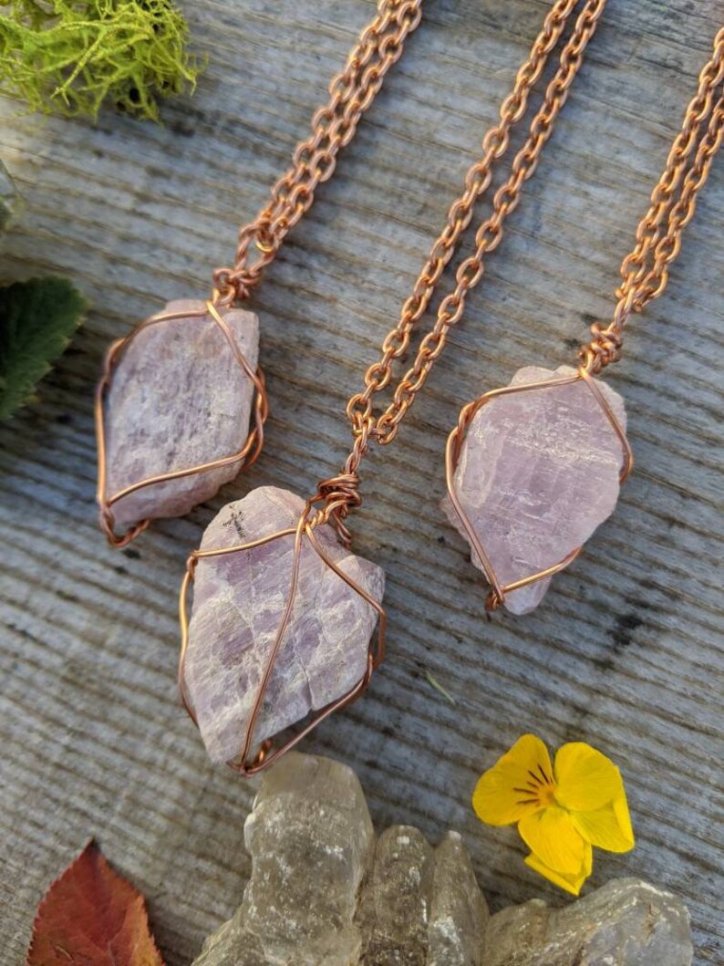 May include: Three delicate necklaces with pink crystal pendants wrapped in copper wire. The necklaces are displayed on a wooden surface with a yellow flower and a brown leaf.